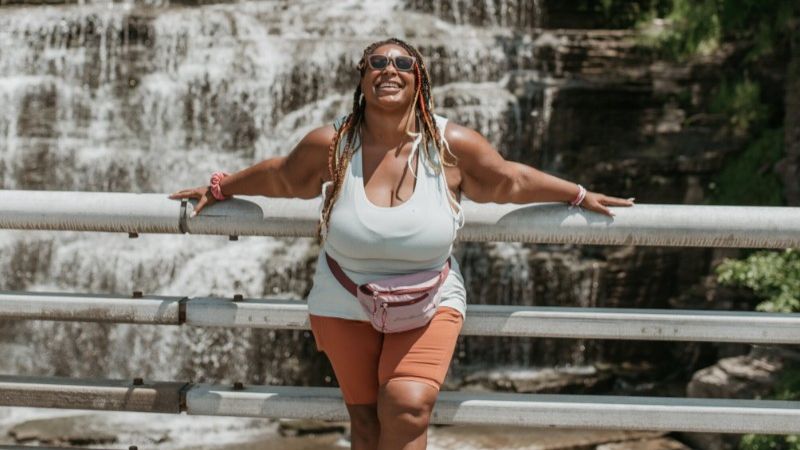 A happy woman wearing a tank top and shorts standing in front of a waterfall