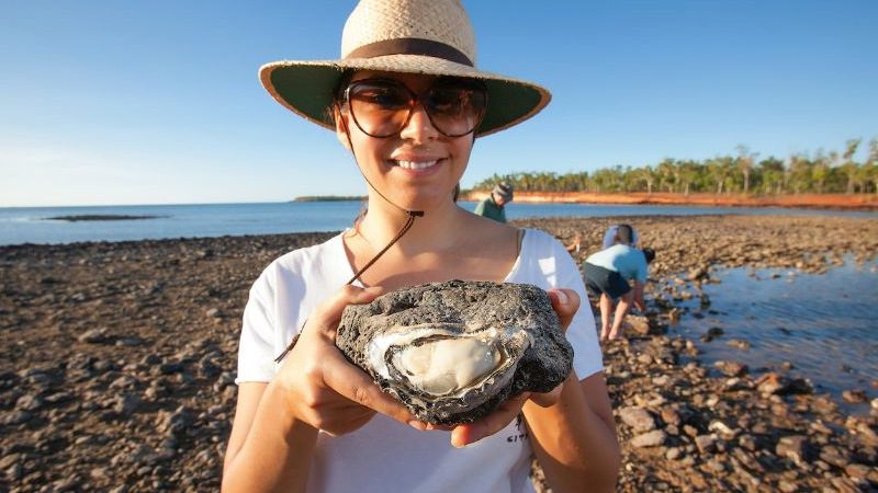 A woman wearing a hat and glasses holds a huge oyster up to the camera