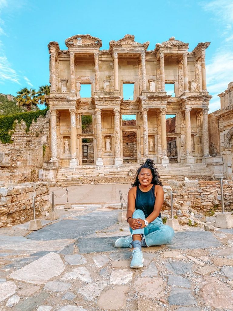 A girl wearing a black top and jeans sitting on the ground in front of an ancient ruin in Turkey