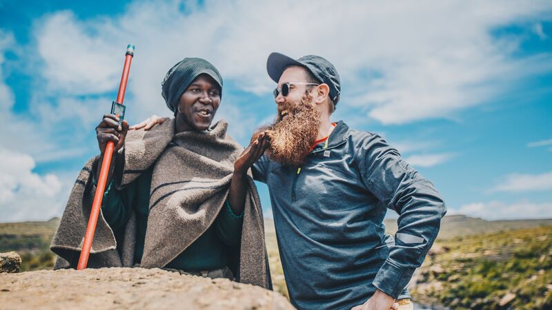 A man with a cap and a beard stands with a man wrapped in a blanket, holding a stick, in South Africa
