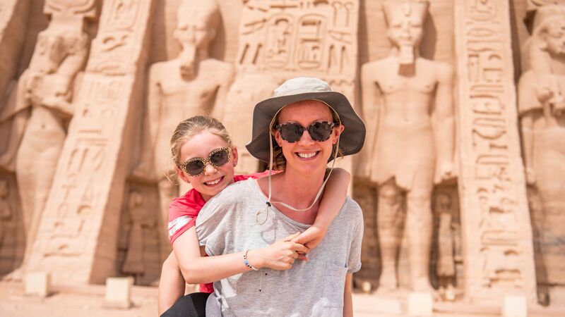 A woman wearing a hat and sunglasses giving a young girl a piggyback in front of a temple in Egypt