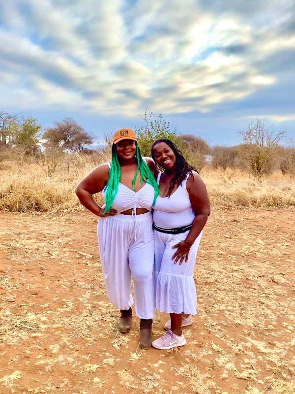 Two women dressed in white, one with an orange cap and long green hair, the other with long dreadlocks, smile for the camera