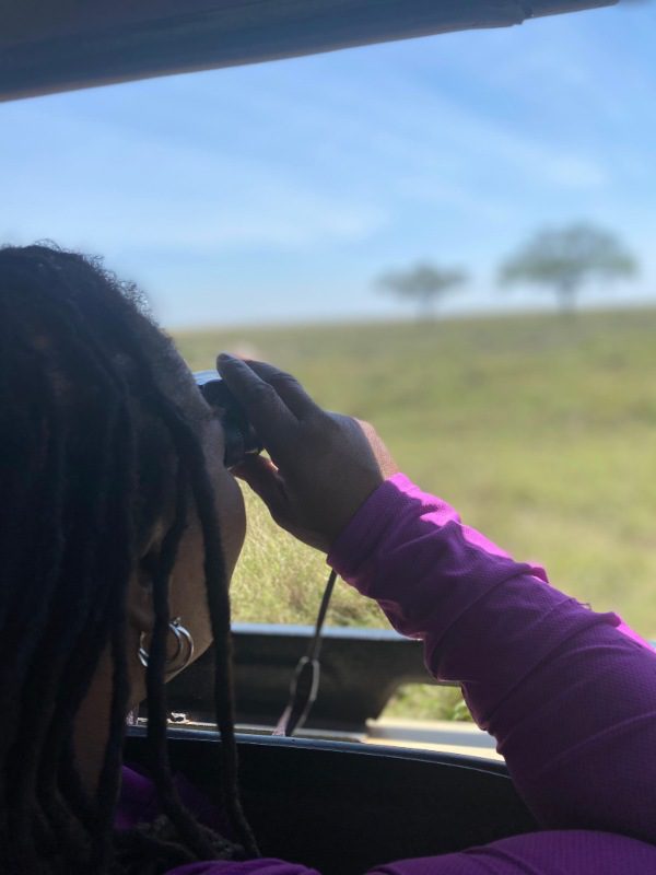 Woman with dreadlocks and purple cat looking through binoculars on safari