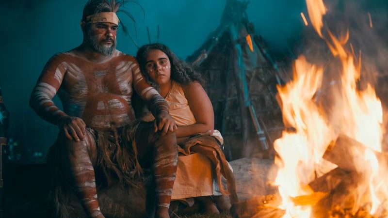 Two Aboriginal actors in traditional dress sit in front of a fire
