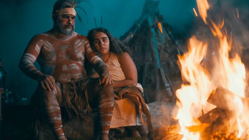 Two indigenous representatives in traditional costume sit in front of a fire