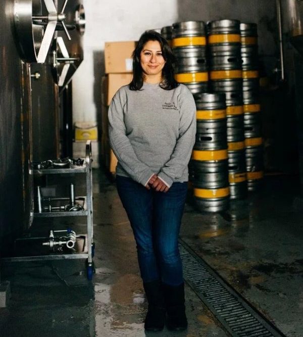 A woman with shoulder-length brown hair wearing jeans and a grey jumper stands in a brewery. There are beer kegs behind her.