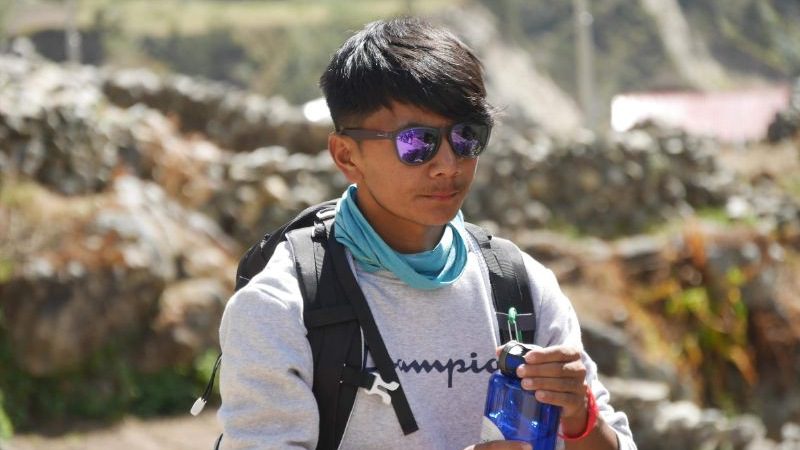 A mountain guide wearing a backpack and sunglasses holds a bottle of water