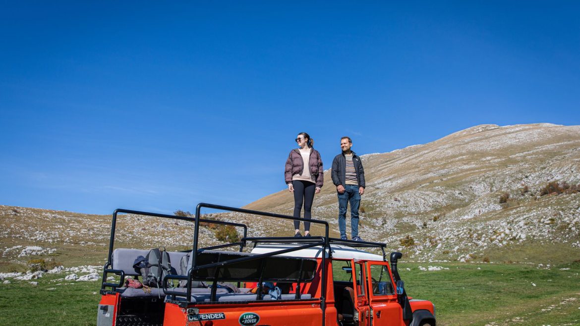 Two people stand on top a safari vehicle with mountains behind