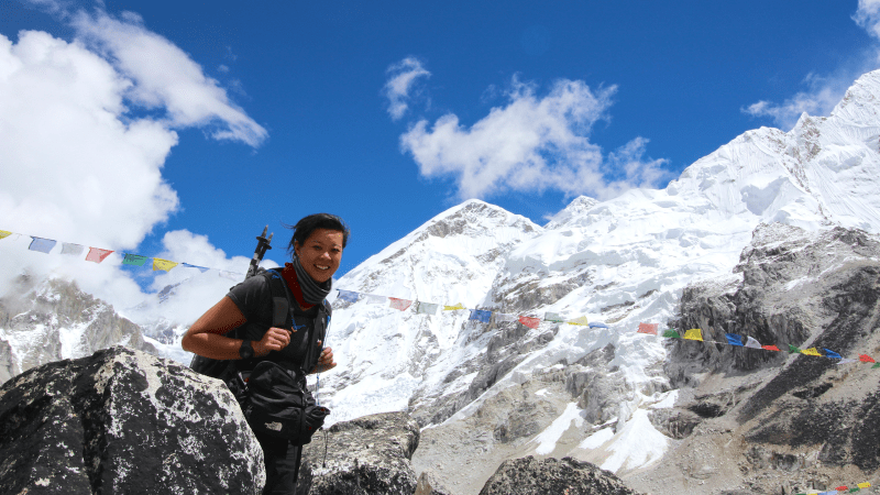A smiling woman backdropped by blue skies, fluttering prayer flags and snowcapped Himalayan mountains