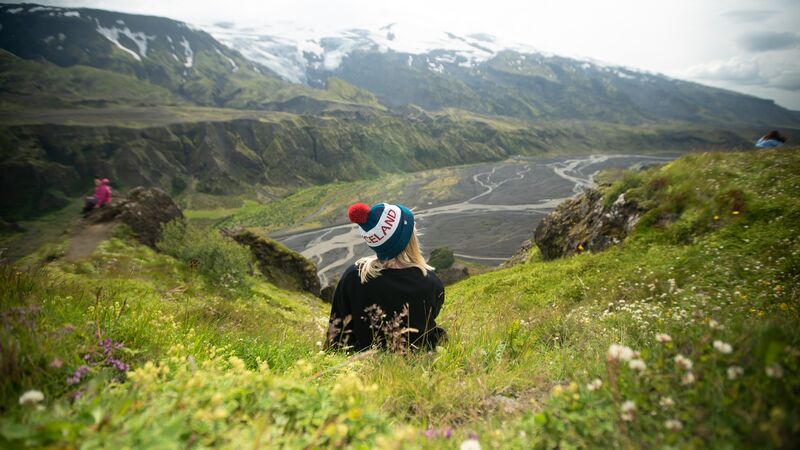 A solo traveler sits on a bright green hillside wearing a hat that says Iceland.