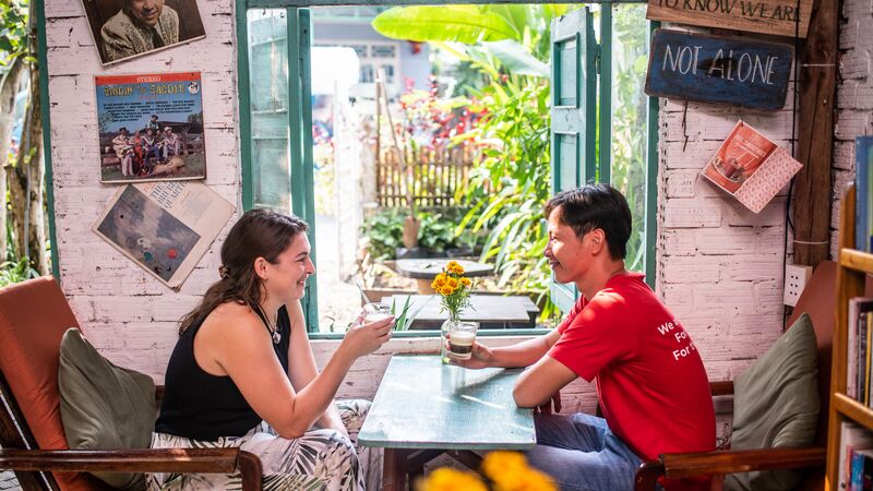 Two people sit across from each other at a table in a colorful coffee shop. Each holds a cup of coffee ready to cheers.