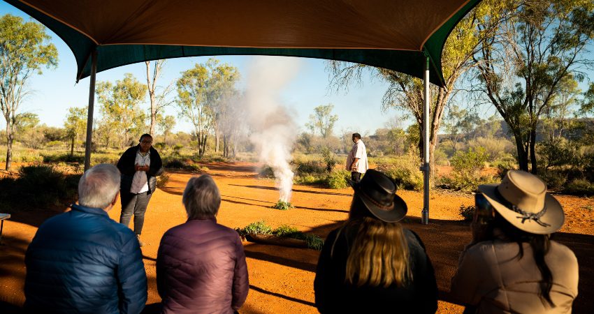 Four travellers sat on a bench while two Aborignal guides explain what a smoking ceremony is