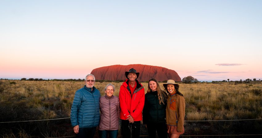 A group of four travellers and their Intrepid leader posing for a photo with Uluru in the background at sunset