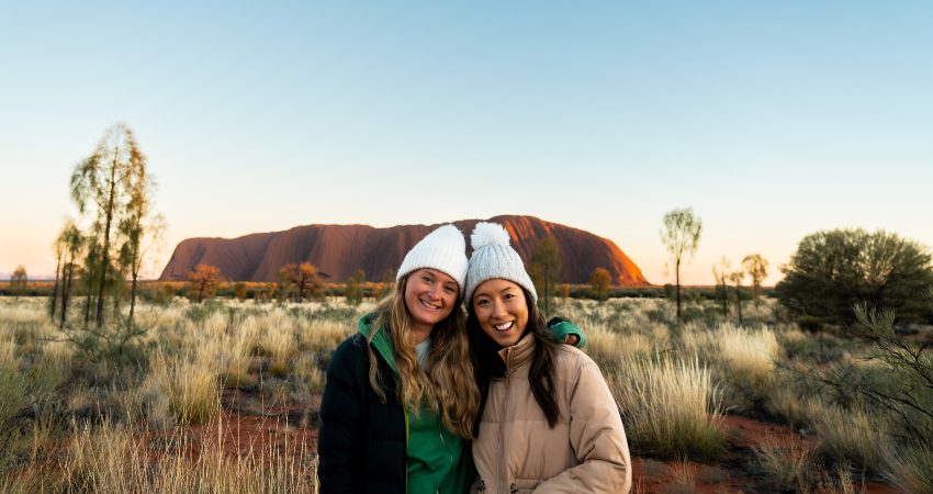 Two travellers smiling with Uluru in the background at sunrise 