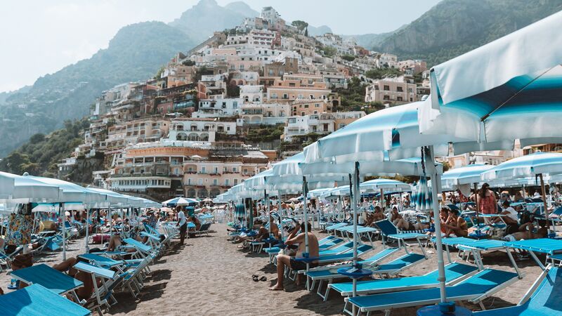 A crowded beach club on the sands of Positano with houses dotting the mountainside in the background. 