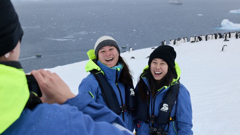 Two travellers posing for a photo on the Antarctic Peninsula with penguins in the background. 