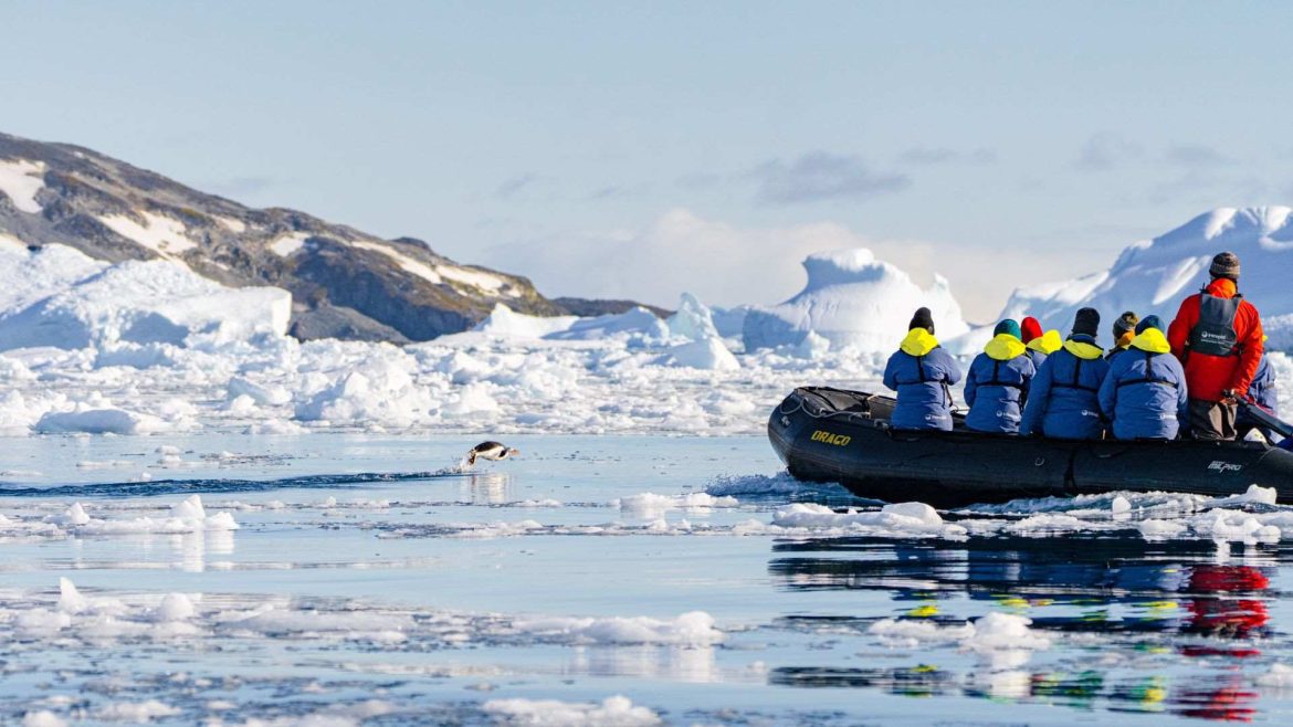 a zodiac filled with a small group of travellers who are looking at a penguin flipping out of the water