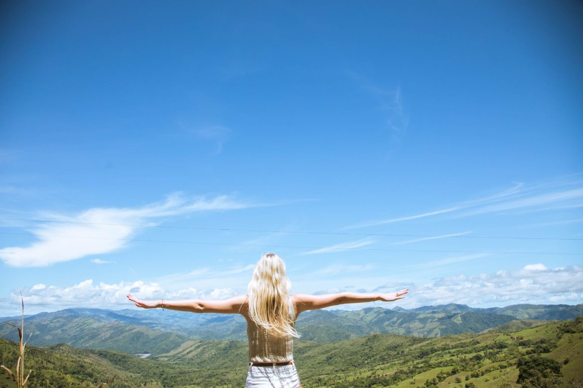 A woman stands facing away from the camera with arms outstretched to her sides as she overlooks a hilly vista in Costa Rica