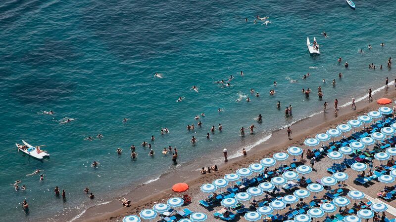 The turquoise waters of the Amalfi Coast with people swimming and lounging under blue and white umbrellas on the pebbly beach.
