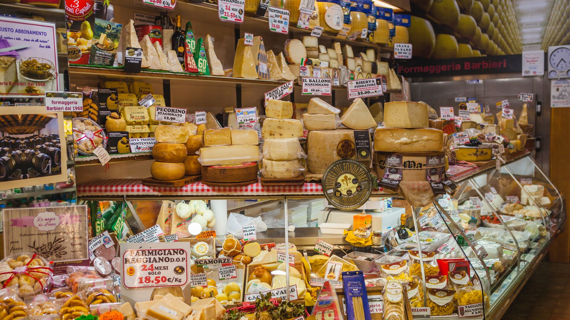 A shop display is stuffed to the brim with different types of cheeses. Black and red signs advertise the name and cost of the cheese.