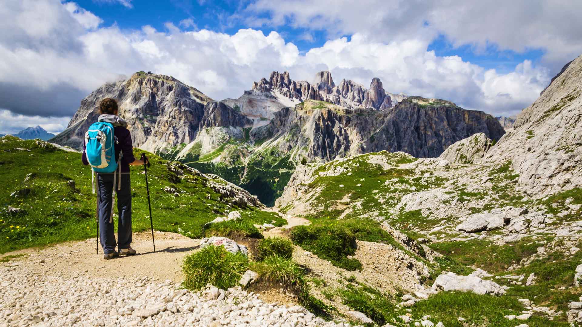 A lone hiker holding a set of poles stands in the middle of a trail taking in a view of the dramatic peaks of the Dolomites in the distance.