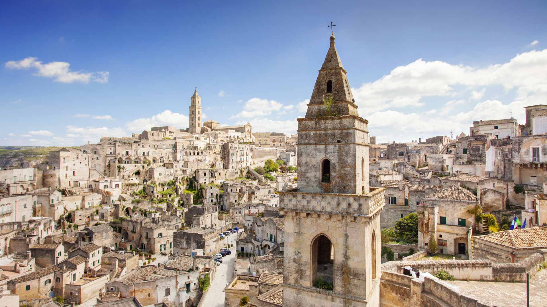 The old town section of Matera is a jumbled puzzle of stacked homes and businesses built into the side of a plateau. The town is a uniform light brown dotted with bright green trees and the occasional flag.