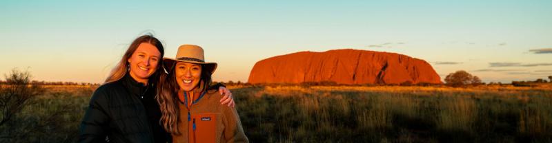 Two travellers posing for a photo in front of Uluru at sunset