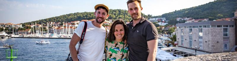 A mixed trio of travellers smiling happily for a photo with a port in the background. 