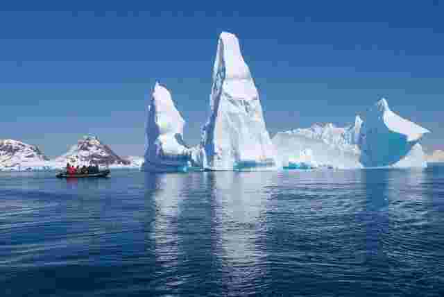 A small zodiac boat floating next to the large formation of a white iceberg. 