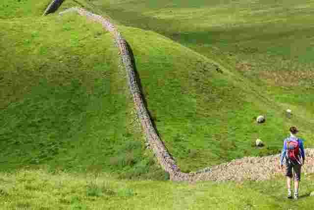 Lone traveller walking along Hadrian's Wall with rolling green hills on either side.