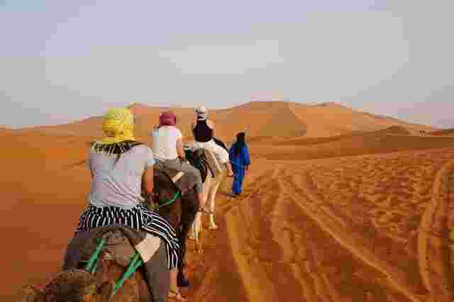 A trail of people riding camels on the orange-golden sand of the Saraha Desert.