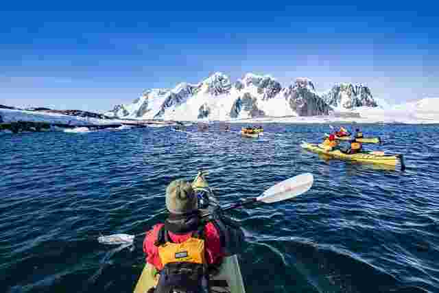 A group of travellers kayaking over the icy waters of Antarctica with iceberg formations in the background.
