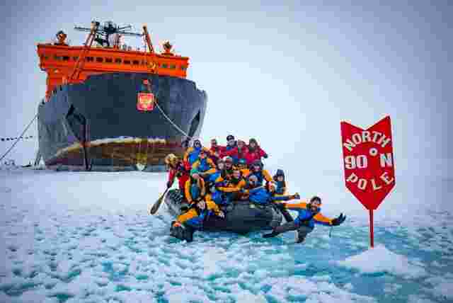 A group of travellers on a zodiac boat at the North Pole 90 degrees sign with the expedition ship in the background.