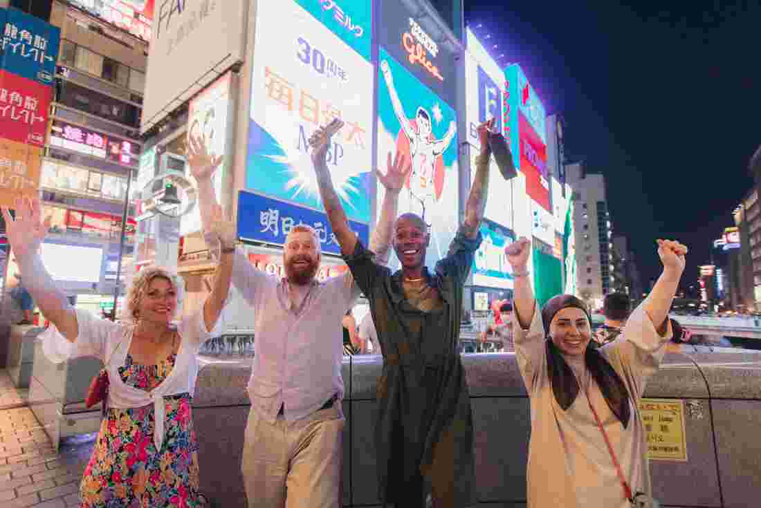Intrepid travellers throw their hands up surrounded by screens and lights in Dotombori