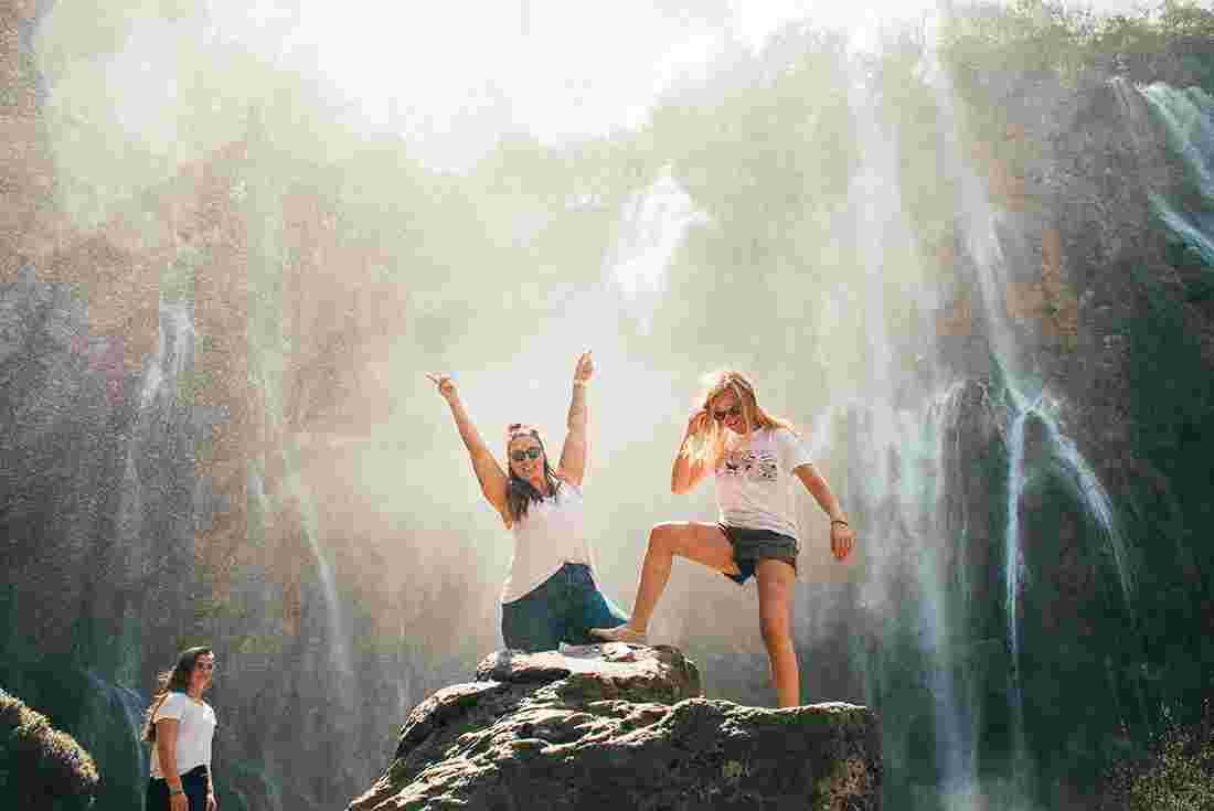 Travellers stand on rock under the spray of waterfall