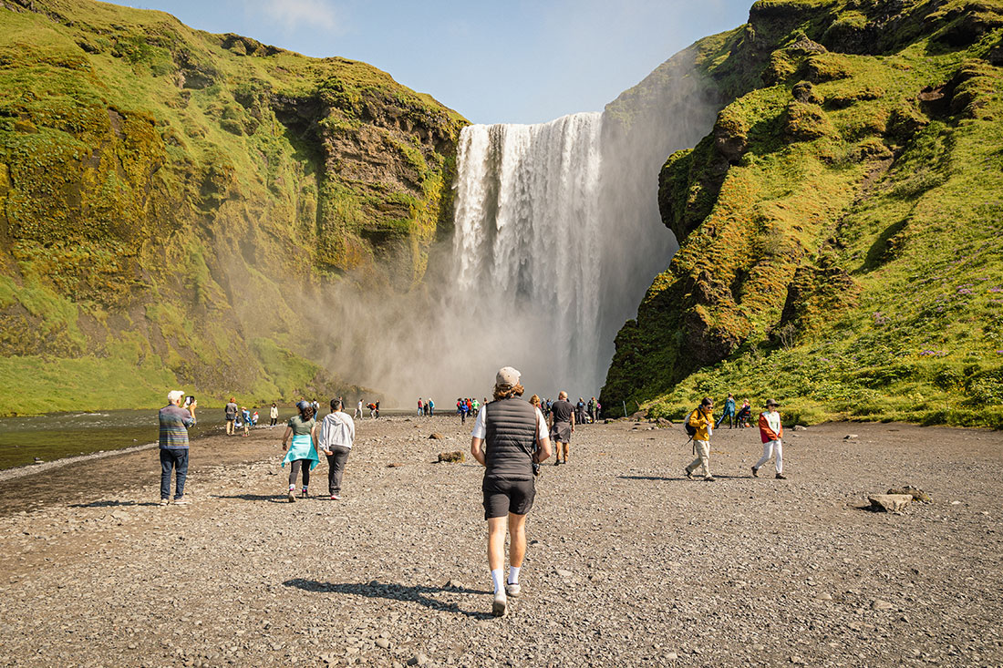 Intrepid traveller walks up to Skogafoss waterfall at the heigh of Iceland summer with numerous other travellers beyond