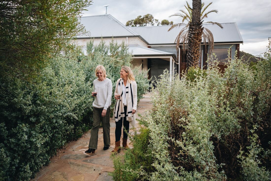 Travellers smiling and enjoying a walk with drinks on the accomodation grounds with green plants around them, Arkaba, South Australia