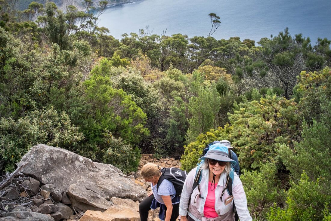 Travellers smiling as they hike up to the lookout, with green trees and the water below behind them, Maria Island, Australia