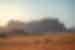 Spire mountain range in distance with two trucks driving past in the distance in an orange sand desert