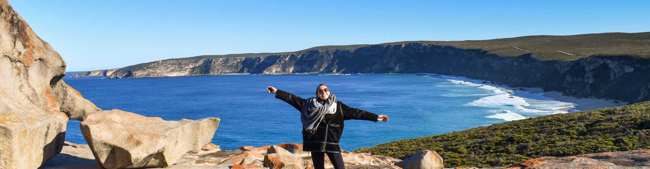 Traveller smiles at camera with arms outstretched looking out over panorama of Kangaroo Island