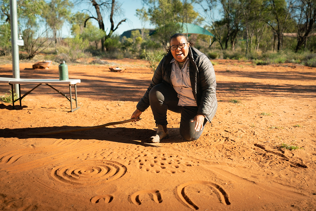 A guide laughs with joy with at the camera kneeling before a set of marks in the sand.