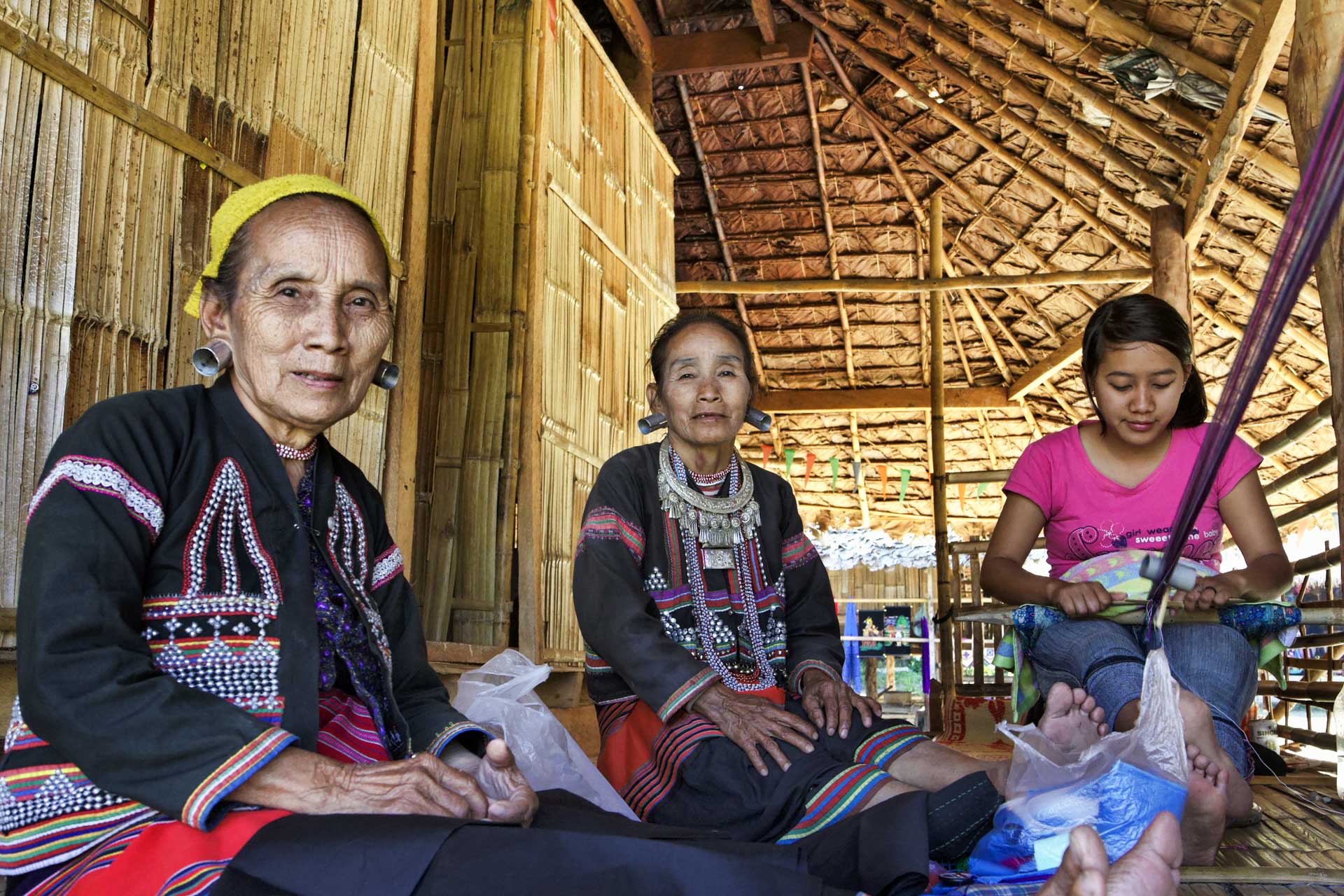 Women of the Ban Huay Pu Keng village, the Karen long neck hill tribe, in traditional dress with their granddaughter weaving alongside